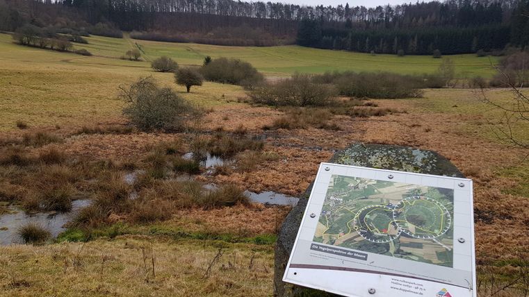 A wide landscape with meadows and a wooded hill in the background. In the foreground, there is an information sign with a map view of the area.