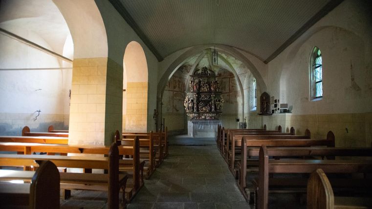 An interior of a church with wooden benches and an elaborate altar setup in the background. The walls are simple, and the windows let in gentle light.