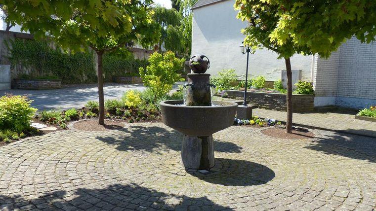 Ein schöner Brunnen steht in einem gepflasterten Innenhof, umgeben von Bäumen und bunten Blumen. Die Umgebung ist ruhig und einladend.
