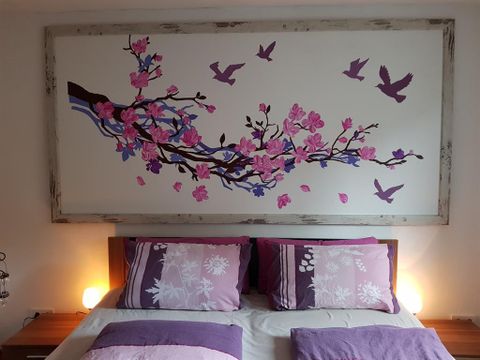 Ein gemütliches Schlafzimmer mit einem großen Wandbild von blühenden Zweigen und Vögeln. Die Bettwäsche ist in lila und rosa Tönen gehalten, mit sanfter Beleuchtung neben dem Bett.