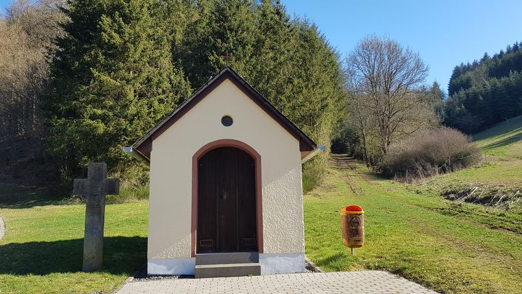 Eine kleine Kapelle steht in einer grünen Hügellandschaft. Im Hintergrund sind Bäume und ein blauer Himmel zu sehen.