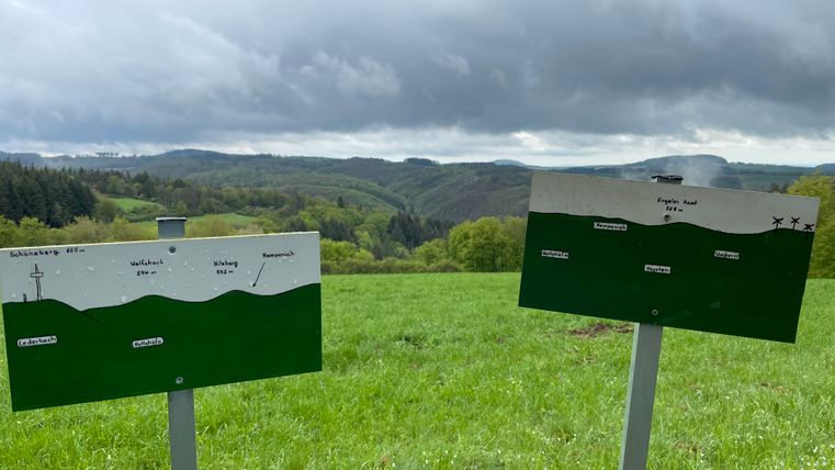 Two signs on a meadow show the names and heights of mountains in the Eifel. Wooded hills and a cloudy sky can be seen in the background.