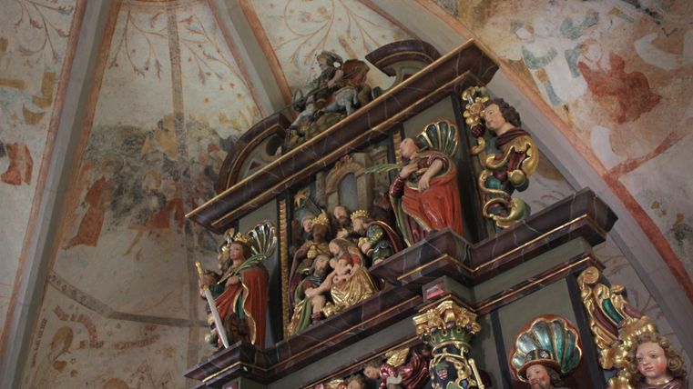 Ein kunstvoll gestalteter Altar mit farbenfrohen Figuren und reichhaltigen Verzierungen. Im Hintergrund sind beeindruckende Wandmalereien zu sehen.