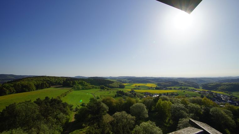Eine weite Landschaft mit grünen Feldern und Wäldern unter einem klaren blauen Himmel. Die Sonne scheint hell über die sanften Hügel.
