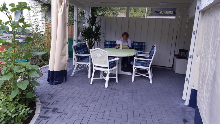 Ein überdachter Außensitzbereich mit einem runden Tisch und Stühlen. Im Hintergrund sitzt eine Person und es sind Pflanzen im Vordergrund zu sehen.