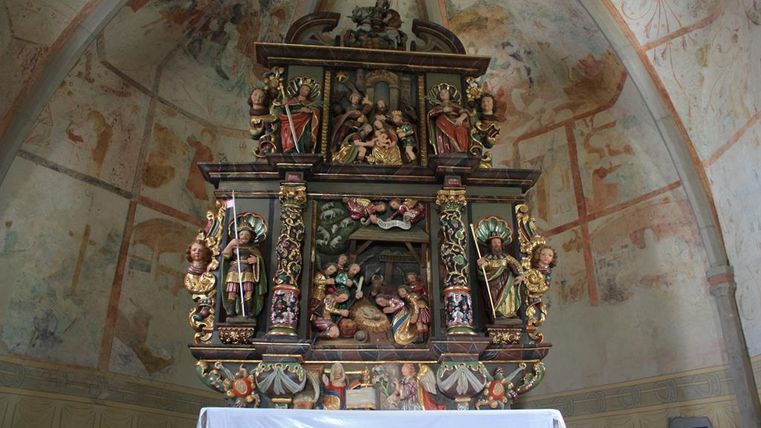 An ornate Baroque altar structure with sculpted figures and detailed decorations. The wall is adorned with colored frescoes that enhance the religious atmosphere.
