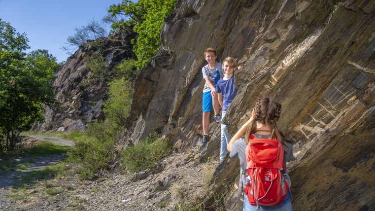 Two children climb on a rock while a person with a red rucksack takes a photo.