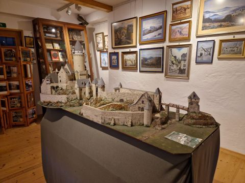 A detailed model view of a castle is on a table. In the background, picture frames can be seen on the wall.