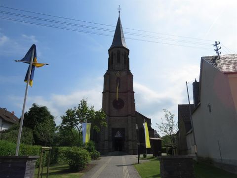 Eine Kirche mit einem hohen Turm steht im Mittelpunkt des Bildes. Umgeben von Grünflächen und flaggenförmigen Dekorationen vermittelt die Szene eine friedliche Atmosphäre.