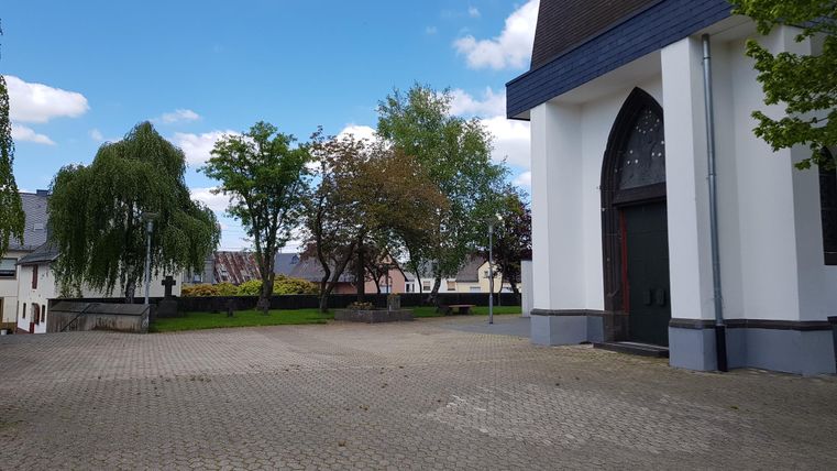 A quiet spot in front of a church with a paved floor. In the background, there are trees and a blue sky.