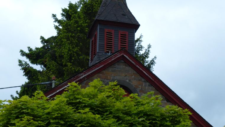 A part of a building with a pointed roof and a small tower can be seen. Green trees and bushes grow around the building.