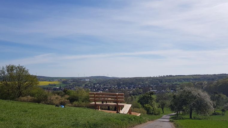 A picturesque landscape with gentle hills and blue sky. In the foreground stands a wooden building, surrounded by meadows and trees.