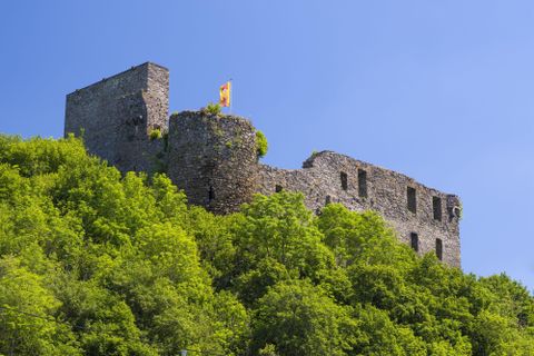 Eine alte Ruine auf einem bewaldeten Hügel. Der Himmel ist blau und sonnig.