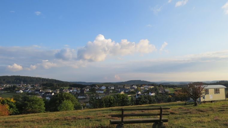 A tranquil landscape with a bench in the foreground and a view of the village in the background. The sky is clear with a few clouds.