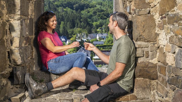 Ein Mann und eine Frau sitzen auf einer Burgmauer und stoßen mit Weingläsern an.