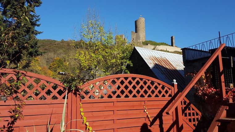 A red garden fence with plants in the foreground. In the background, a castle rises on a hill under a clear blue sky.