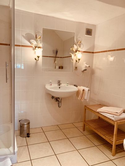 Ein helles Badezimmer mit Fliesenboden und modernen Armaturen. Es gibt eine Dusche, einen Waschbecken mit Spiegel und einen Bogen aus Holzregalen.