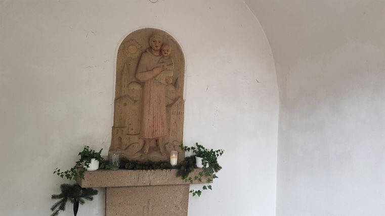 Eine Statue in einer Nische, umgeben von Pflanzen und Kerzen. Die Wand ist schlicht und weiß gestrichen.
