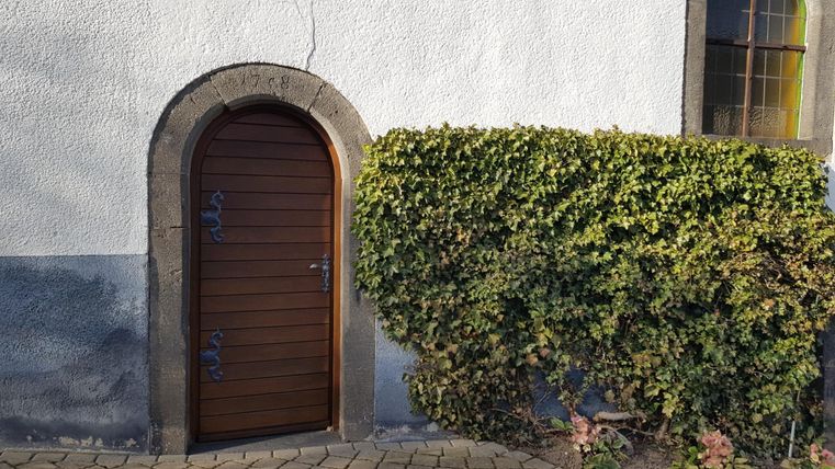 Eine kleine, runde Holztür an einer weißen Wand. Daneben wächst eine dichte, grüne Hecke.
