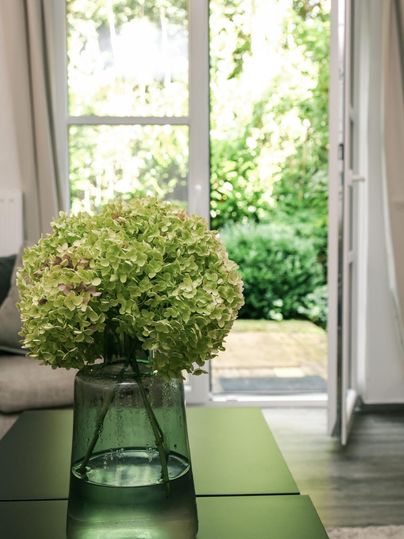 A beautiful bouquet of flowers in a glass vase is on a table. In the background, there is an open door overlooking a green garden.