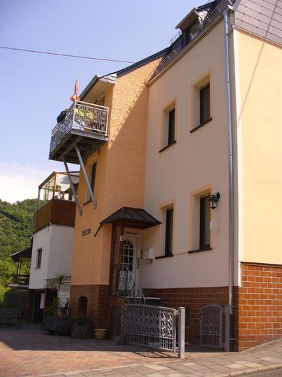 Ein modernes, zweigeschossiges Haus mit einem Balkon und einer Eingangstür. Es ist von Bäumen umgeben und befindet sich in einer ruhigen Umgebung.