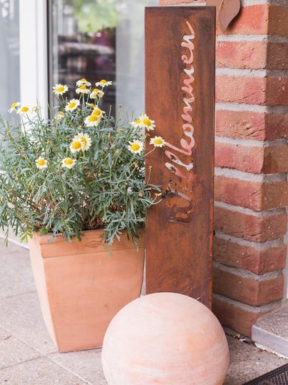A decorative flower pot with a yellow flower stands next to a wooden sign that reads "Welcome." Next to it lies a round stone light on the ground.