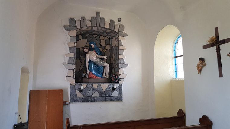 Eine religiöse Statue zeigt Maria mit dem gekreuzigten Jesus in einer Kapelle. Der Raum hat helles Licht und eine einfache, friedliche Atmosphäre.