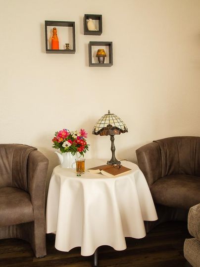 Ein gemütlicher Sitzbereich mit zwei Sessel und einem runden Tisch. Auf dem Tisch stehen eine Lampe, Blumen und ein Getränk.