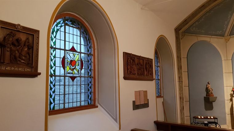An interior of a church with a colorful stained glass window and wooden reliefs on the wall. In the background, a statue can be seen.