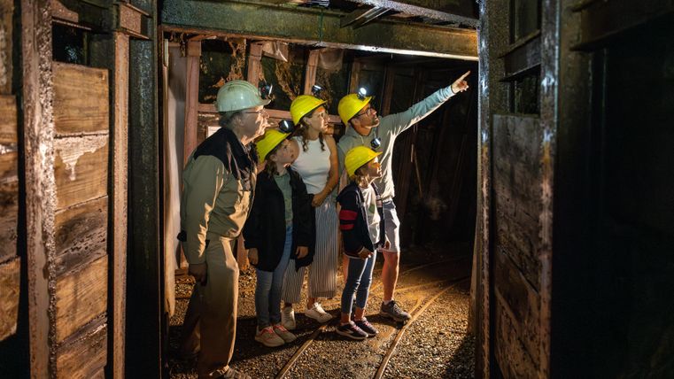 Eine Gruppe von Personen in einem Bergwerk, die Helme tragen und sich umschauen. Eine Person zeigt auf etwas Interessantes.