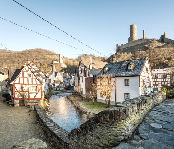 Fachwerkhäuser in Monreal, ein Bach fließt durch das Dorf. Im Hintergrund eine Burgruine auf einem Hügel unter klarem Himmel., © Eifel Tourismus GmbH, D. Ketz