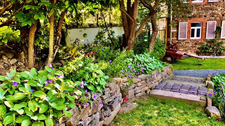 A beautiful garden with colorful flowers and a paved path. In the background, you can see a brick house with windows.