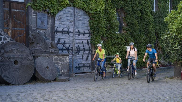 Eine Familie fährt mit Fahrrädern an einem alten Gebäude mit Efeu und einer Löwenstatue vorbei.