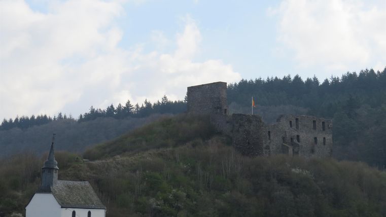 Eine alte Burg thront auf einem bewaldeten Hügel. Unterhalb befindet sich eine kleine weiße Kirche.