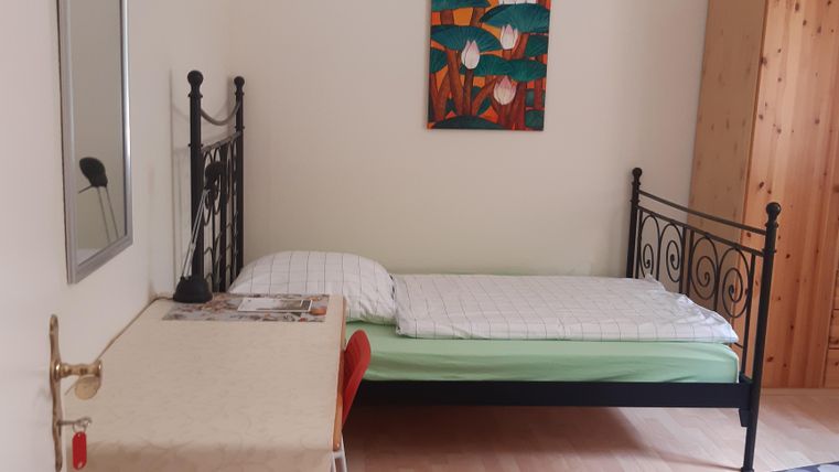 Ein einfaches Zimmer mit einem Einzelbett und einem Holzschrank. Ein Teppich und ein Gemälde sorgen für eine gemütliche Atmosphäre.