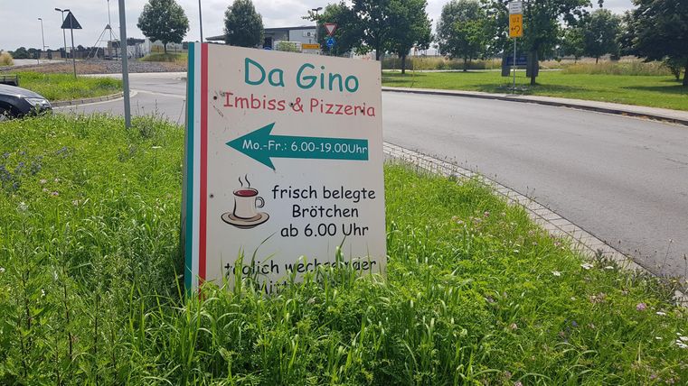 A sign for "Da Gino" with information about filled rolls and pizzas. It shows an arrow pointing left and is surrounded by grass.