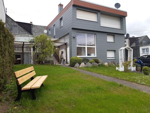 Ein modernes Haus mit grauer Fassade und einem kleinen Garten. Eine Holzbank steht vor dem Haus in einer grünen Wiese.