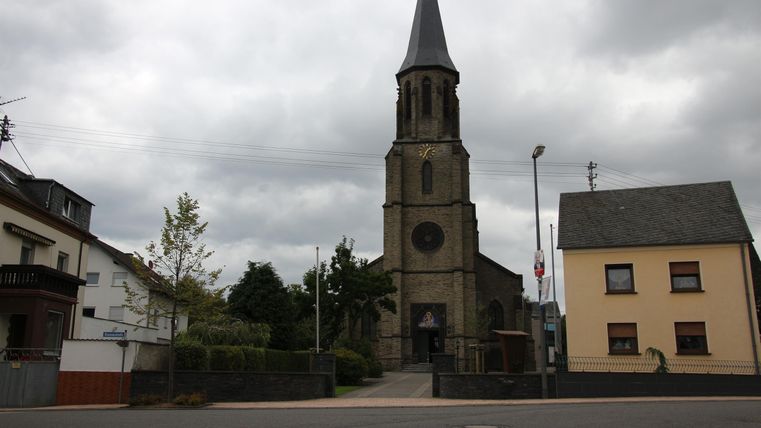 Eine alte Kirche mit einem spitzen Turm steht im Vordergrund. Die Wolken am Himmel verleihen der Szene eine ruhige Atmosphäre.