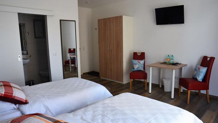 A modern room with two single beds, a table, and two chairs. A television is mounted on the wall, and there is a mirror as well as a wardrobe.