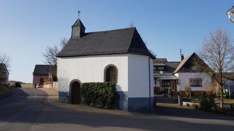 A small white building with a black roof stands on a quiet street. In the background, there are more residential houses and a clear sky.