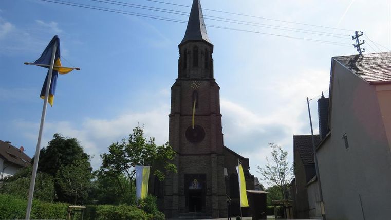 Eine Kirche mit einem hohen Turm und bunten Flaggen im Vordergrund. Der Himmel ist klar und es gibt viel grünes Laub um die Kirche herum.