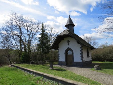 Eine kleine Kapelle steht umgeben von Bäumen und Gras. Der Himmel ist teilweise bewölkt.