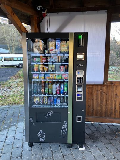Ein Verkaufsautomat mit einer Vielzahl von Snacks und Getränken. Er befindet sich unter einem überdachten Bereich mit Holzverkleidung.