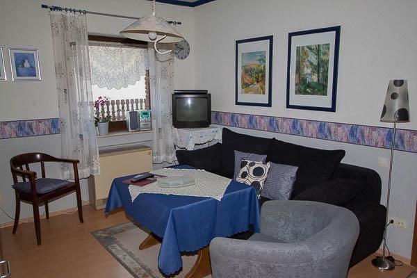 Ein gemütliches Wohnzimmer mit einem dunkelblauen Sofa und einem grauen Sessel. An der Wand hängen Bilder und ein Fernseher steht auf einem Möbelstück.