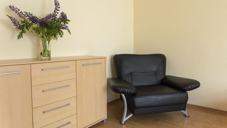 A cozy room with a black armchair and a wooden dresser. A vase with purple flowers is on the table.