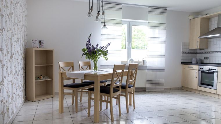 A bright dining room with a table and chairs. A flower vase in the center and modern kitchen cabinets in the background.