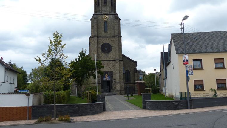 Eine Kirche mit einem hohen Turm steht im Hintergrund, umgeben von Bäumen und Wohnhäusern. Der Himmel ist bewölkt und es ist eine ruhige Straße zu sehen.