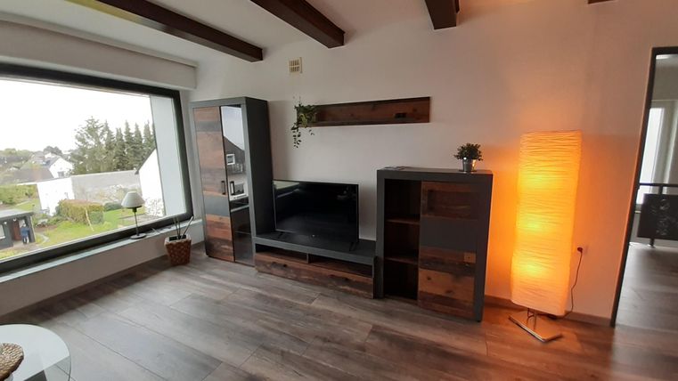 Ein modernes Wohnzimmer mit Holzdeckenbalken und großem Fenster. Der Raum ist minimalistisch eingerichtet mit einem Bücherregal und einer Stehlampe.