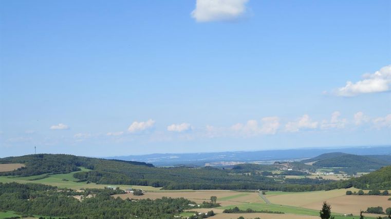 Eine weite Landschaft mit sanften Hügeln und Feldern unter einem klaren blauen Himmel. Im Hintergrund sind einige Bäume und leicht bewaldete Flächen sichtbar.