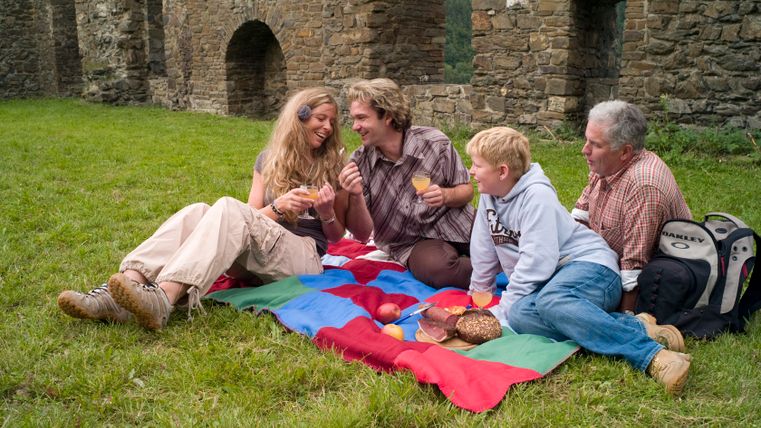 Vier Personen machen ein Picknick auf einer bunten Decke vor einer alten Steinmauer.